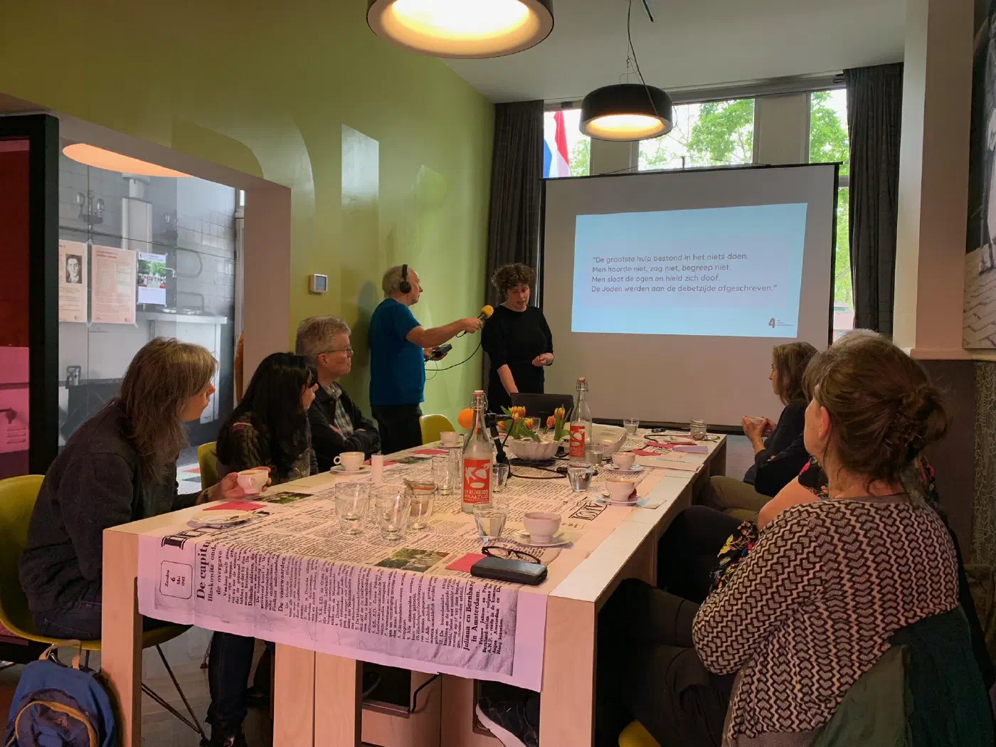 Spreker bij een projectiescherm wordt opgenomen terwijl gasten aan tafel luisteren