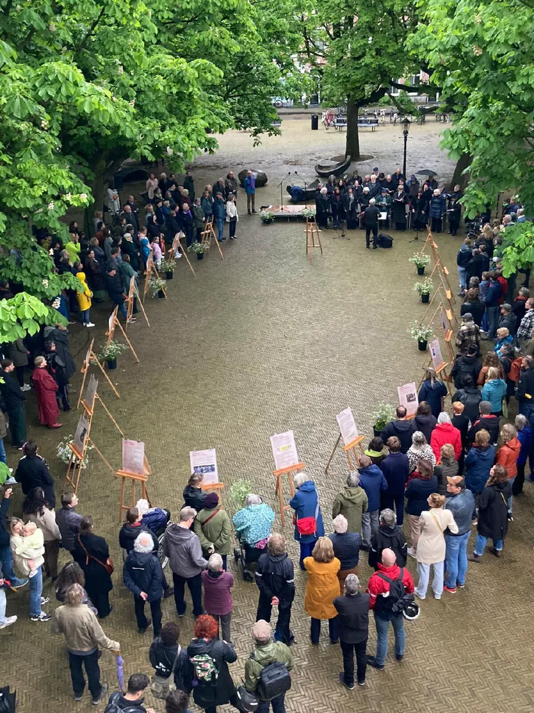 Bovenaanzicht van de herdenking: publiek rond gedenkposters op ezels en een koor