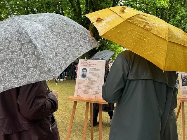 Bezoekers onder paraplu’s lezen een gedenkposter op een ezel in de regen