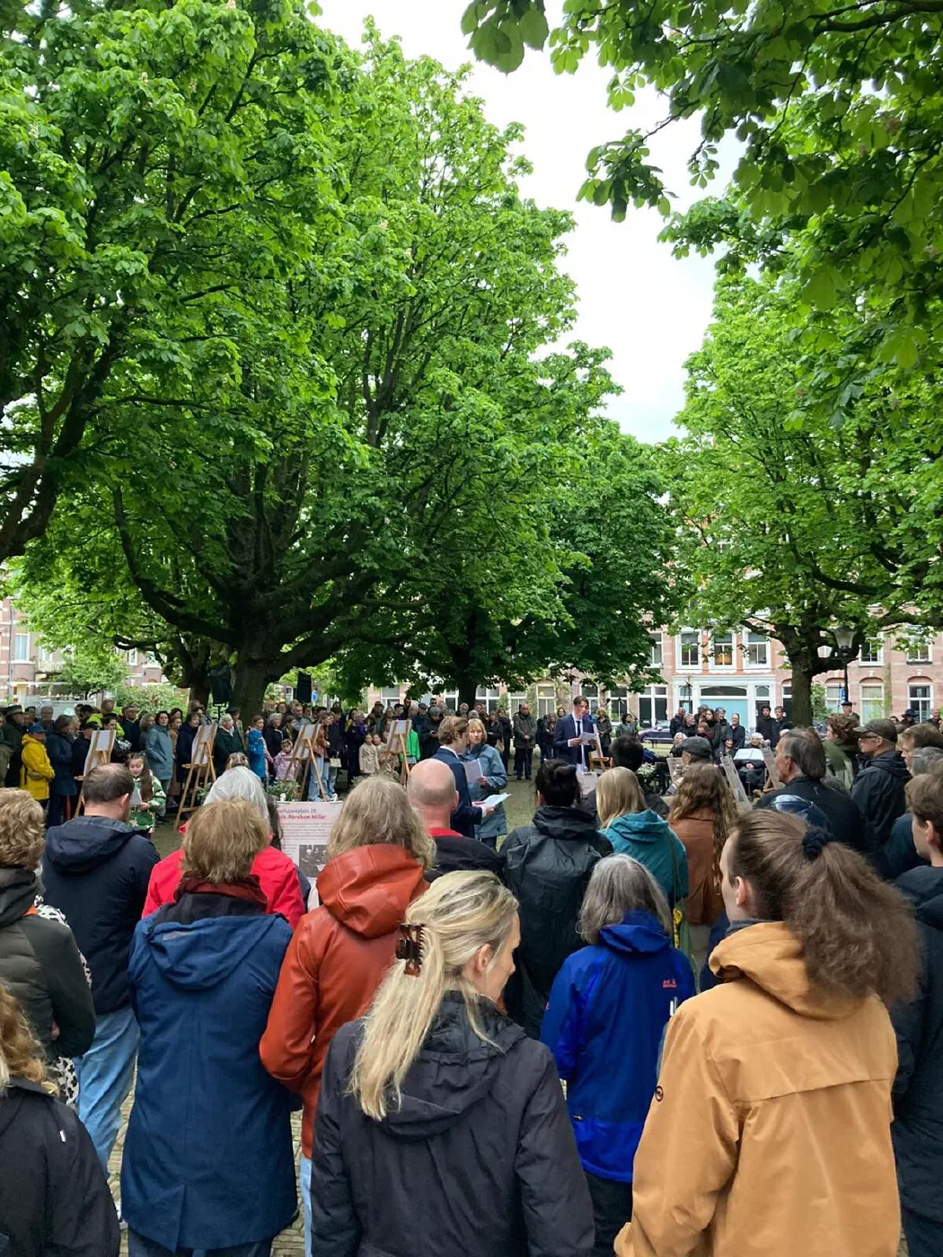 Dicht opeengepakt publiek onder kastanjebomen luistert naar sprekers op het plein