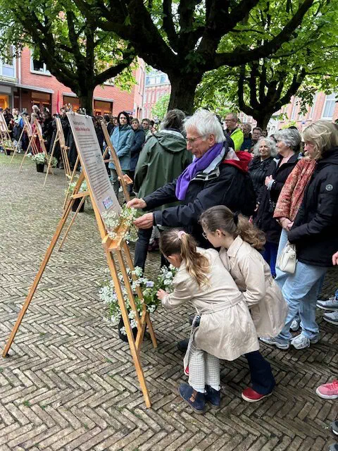 Twee jonge meisjes en een oudere man leggen bloemen bij een gedenkposter