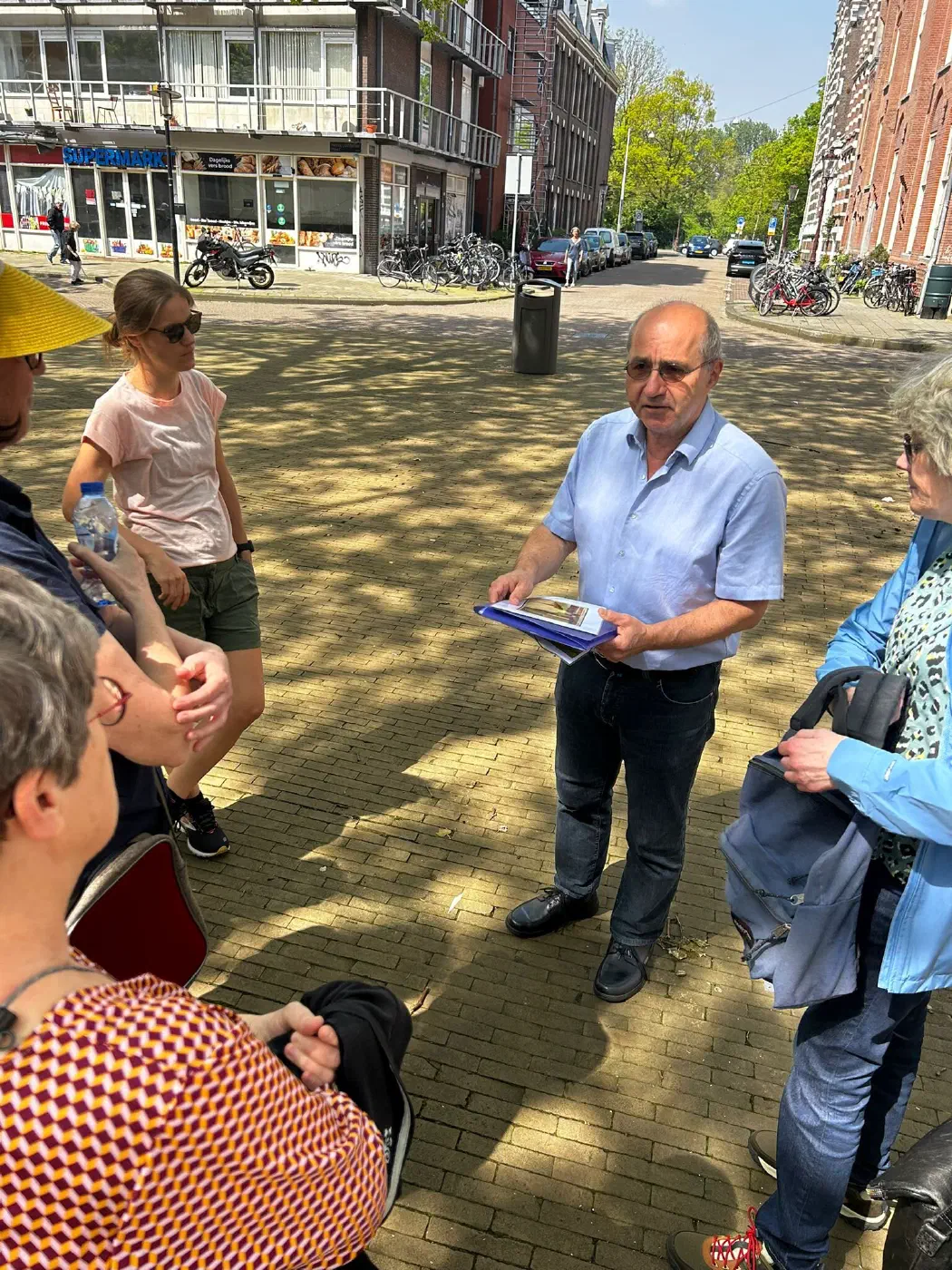 Gids met een map vol foto’s spreekt deelnemers toe op een zonnig plein