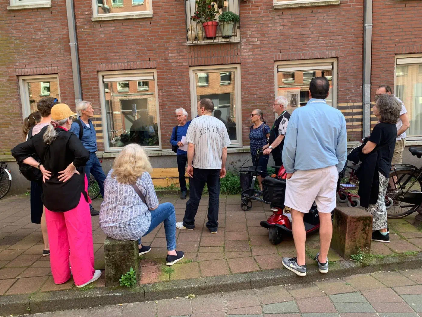 Grote groep deelnemers, onder wie mensen met rollator en scootmobiel, luistert naar de gids