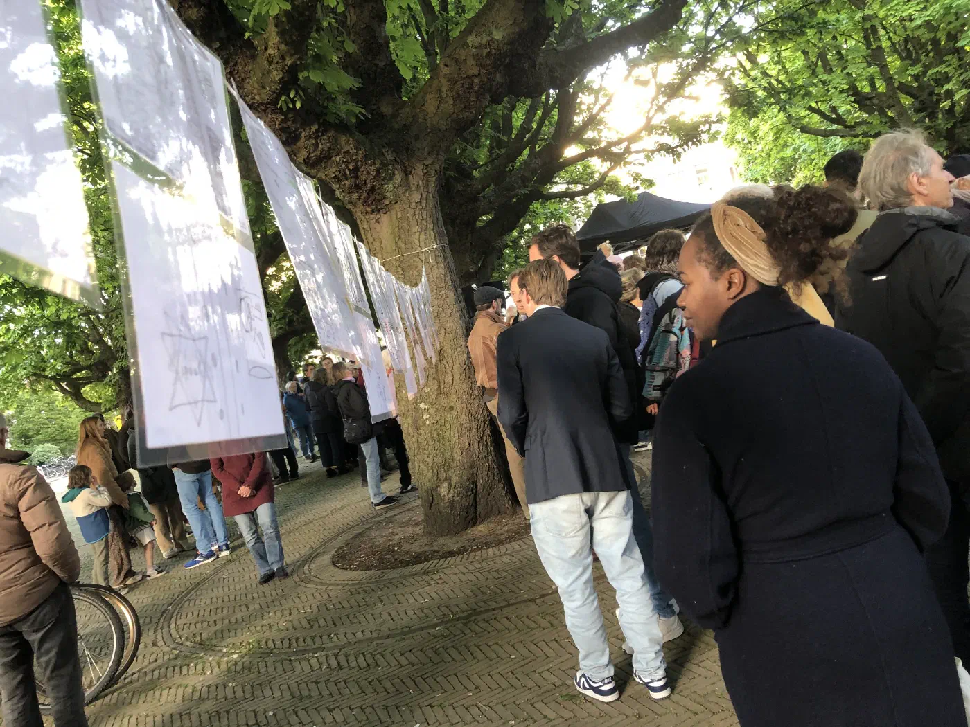 Tekeningen van leerlingen hangen aan een lijn tussen de bomen, bezoekers kijken