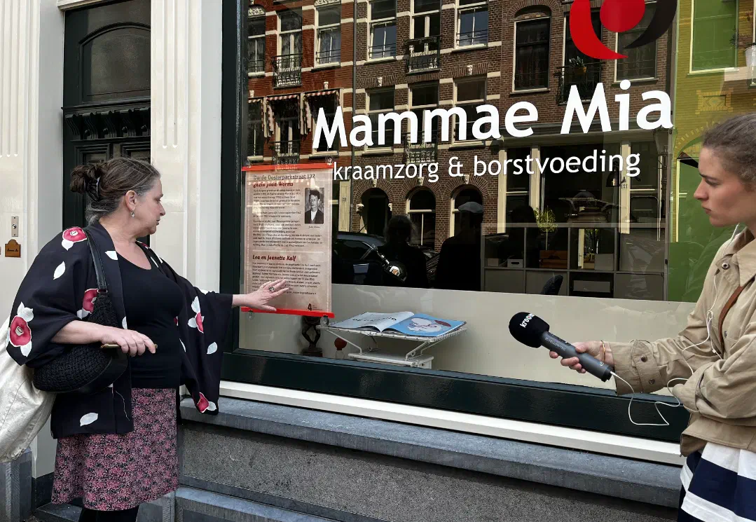 Rondleider wijst op een gedenkposter in een etalage terwijl een verslaggever opneemt