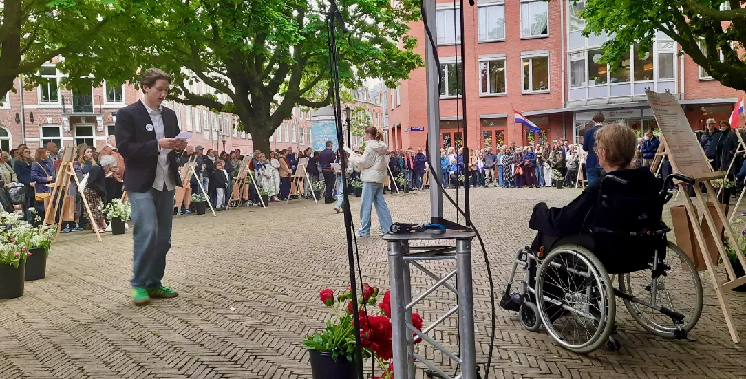 Jonge spreker leest voor, naast een vrouw in een rolstoel, met publiek eromheen