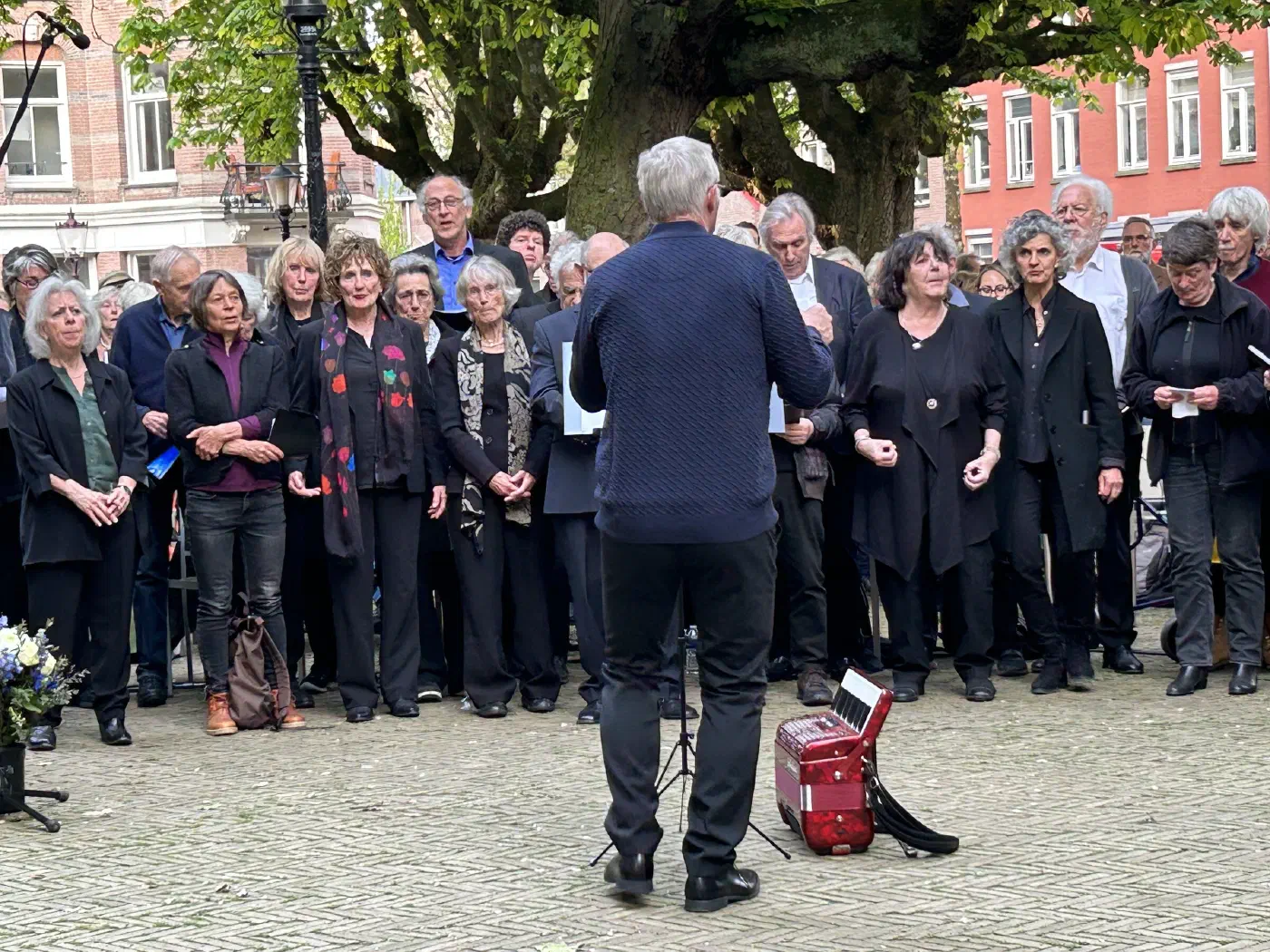 Dirigent met de rug naar de camera voor een zingend koor onder de kastanjebomen