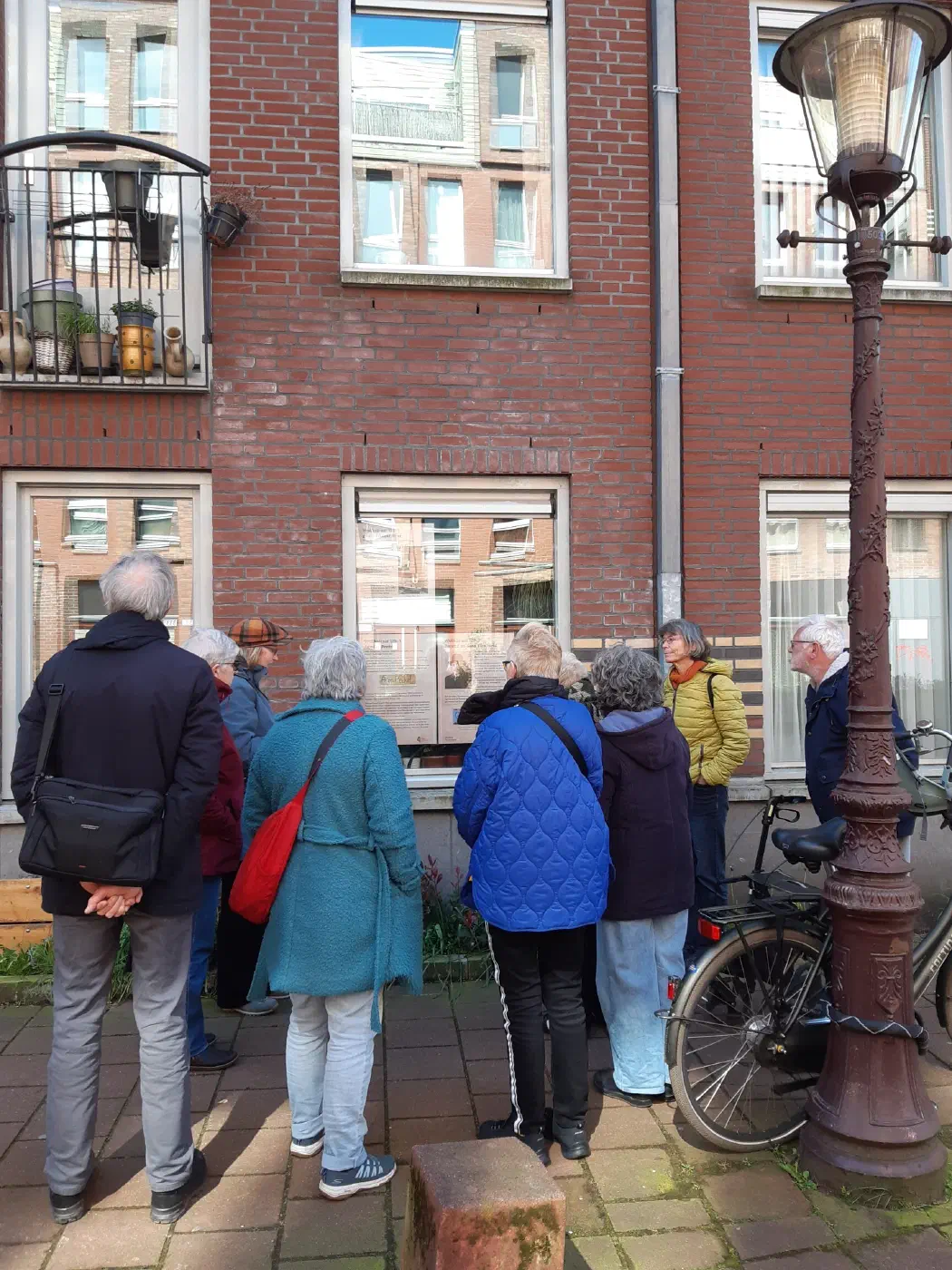 Groep buurtbewoners leest gedenkposters achter het raam van een bakstenen huis