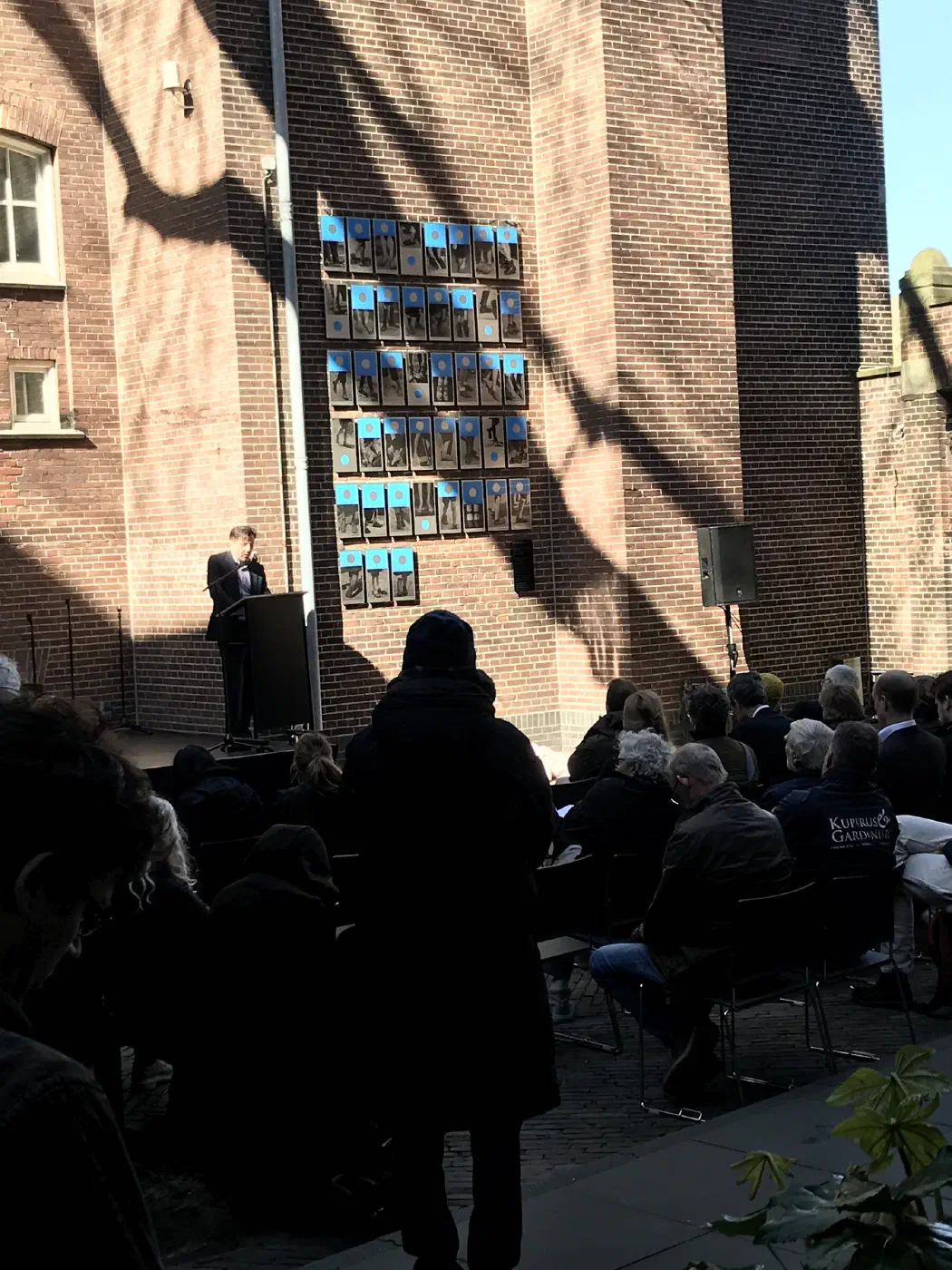 Spreker bij het vernieuwde monument met 46 foto’s van voeten op de schoolmuur