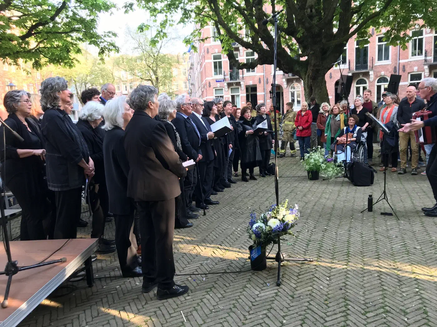 Koor in het zwart zingt onder de kastanjeboom, omringd door publiek en bloemen