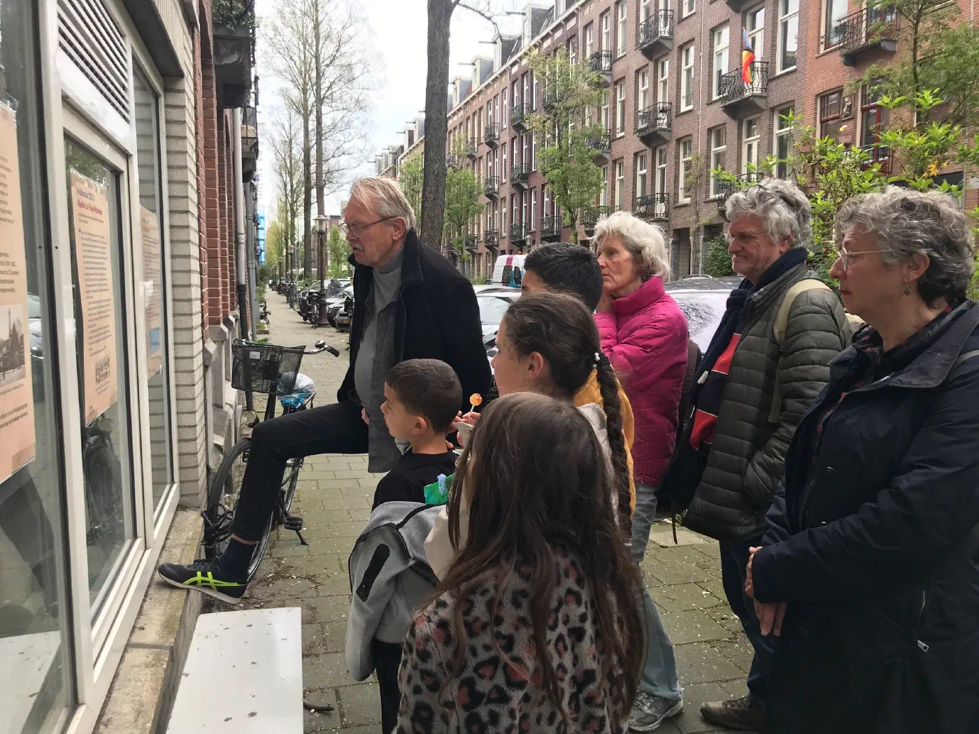 Volwassenen en kinderen lezen samen gedenkposters achter een raam in de Vrolikstraat